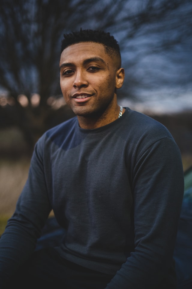 Male with dark hair and brown eyes, wearing a blue sweater and a silver chain posing in front of a tree