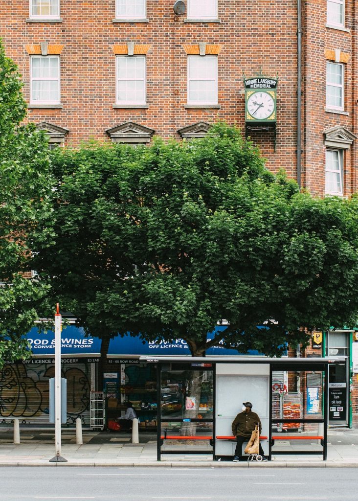 A picture of a bus stop and a man waiting in front of it wearing a covid mask. Tree and shops behind him