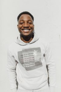 Black male with dark hair, smiling with white teeth, he is very happy. He is wearing a white jumper