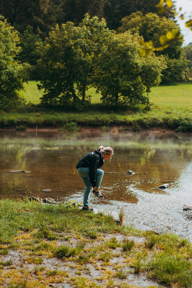 Picture of a blonde female out in nature. Behind her is a lake with stones and trees. She is wearing a NorthFace coat, blue skinny jeans and vans shoes.