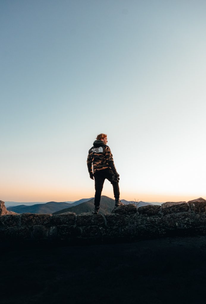 Picture of a man standing before a beautiful landscape, with a series of mountains at sunset. He is strawberry blonde wearing a camouflage NorthFace jumper.