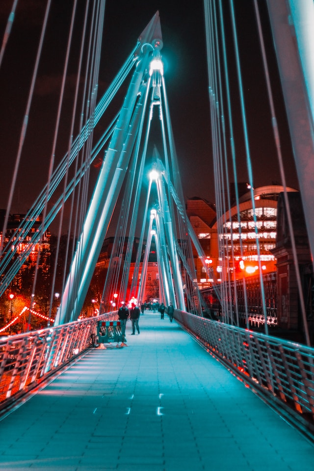 A picture of a bridge with people walking across it. red ad white lights illuminate the skyline, apartments in the background
